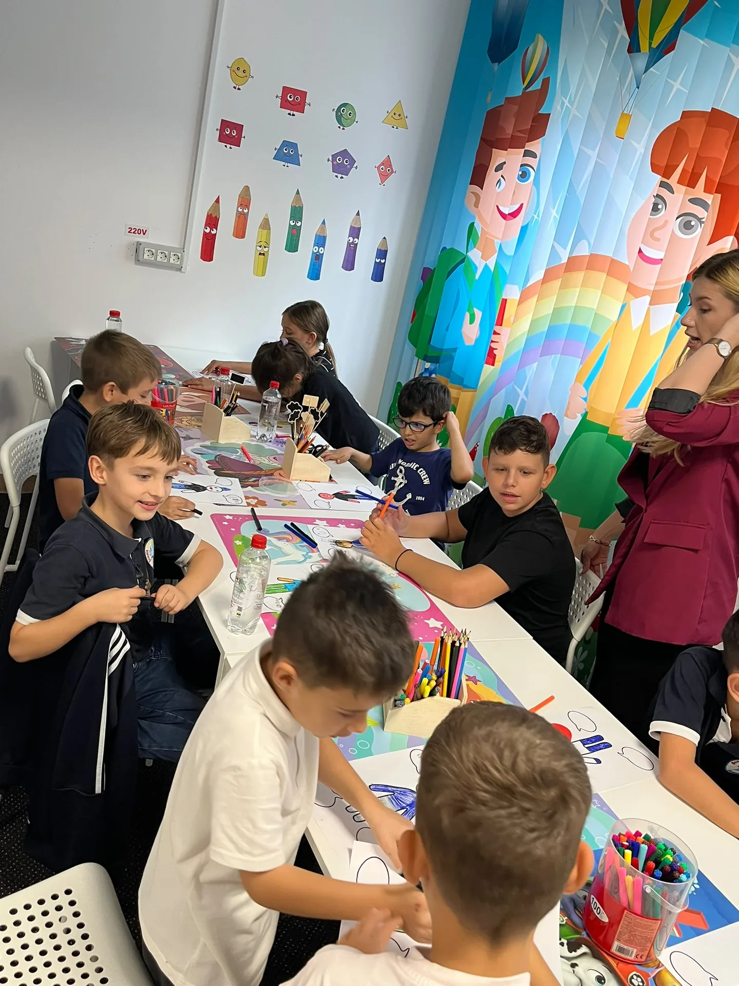 Copiii lucrează cu carioci la o masă lungă, lângă perdele ilustrate cu personaje și un curcubeu.