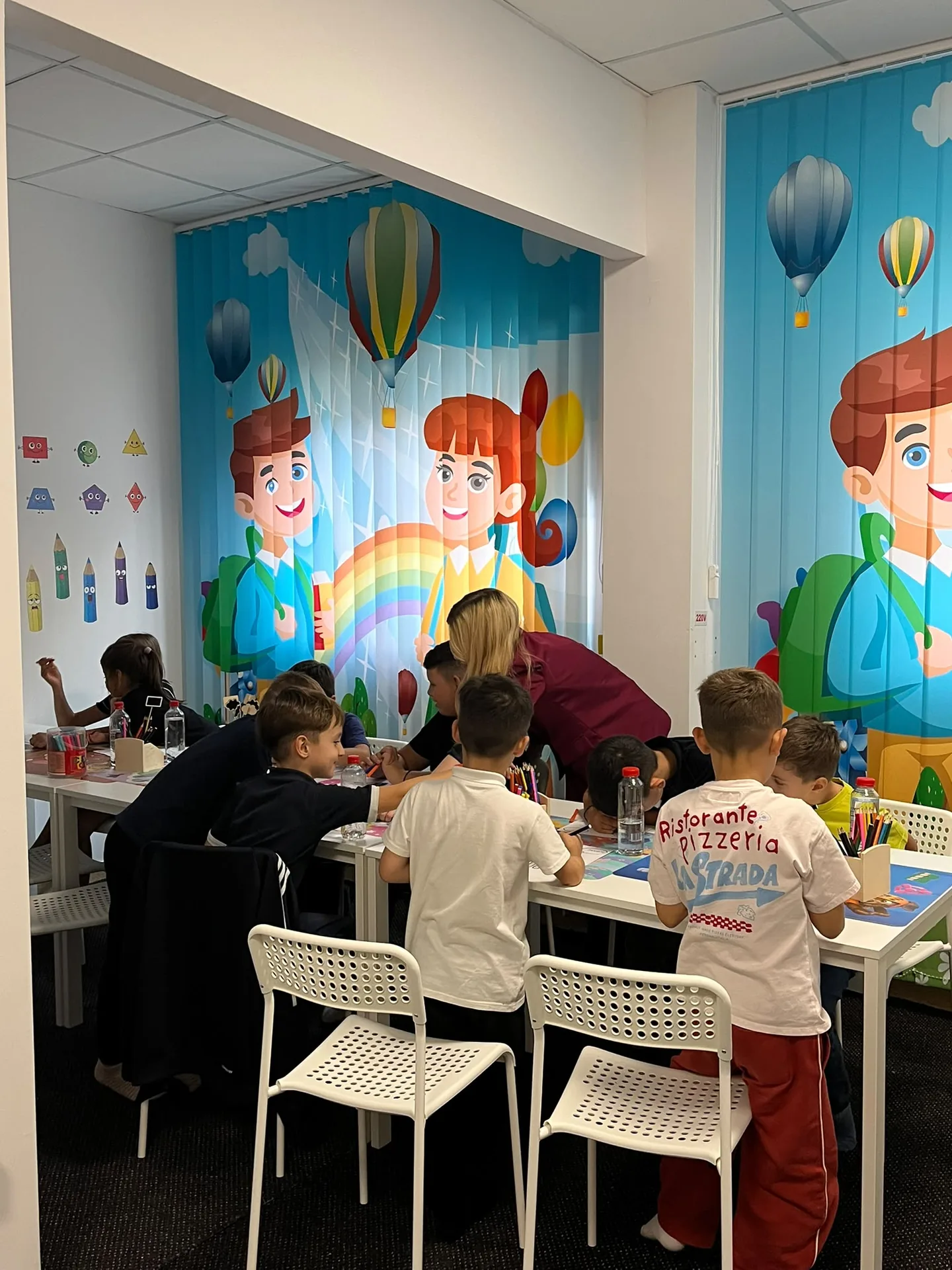 Copiii colorează în grup, iar o adultă se apleacă lângă ei la masa cu creioane și carioci.