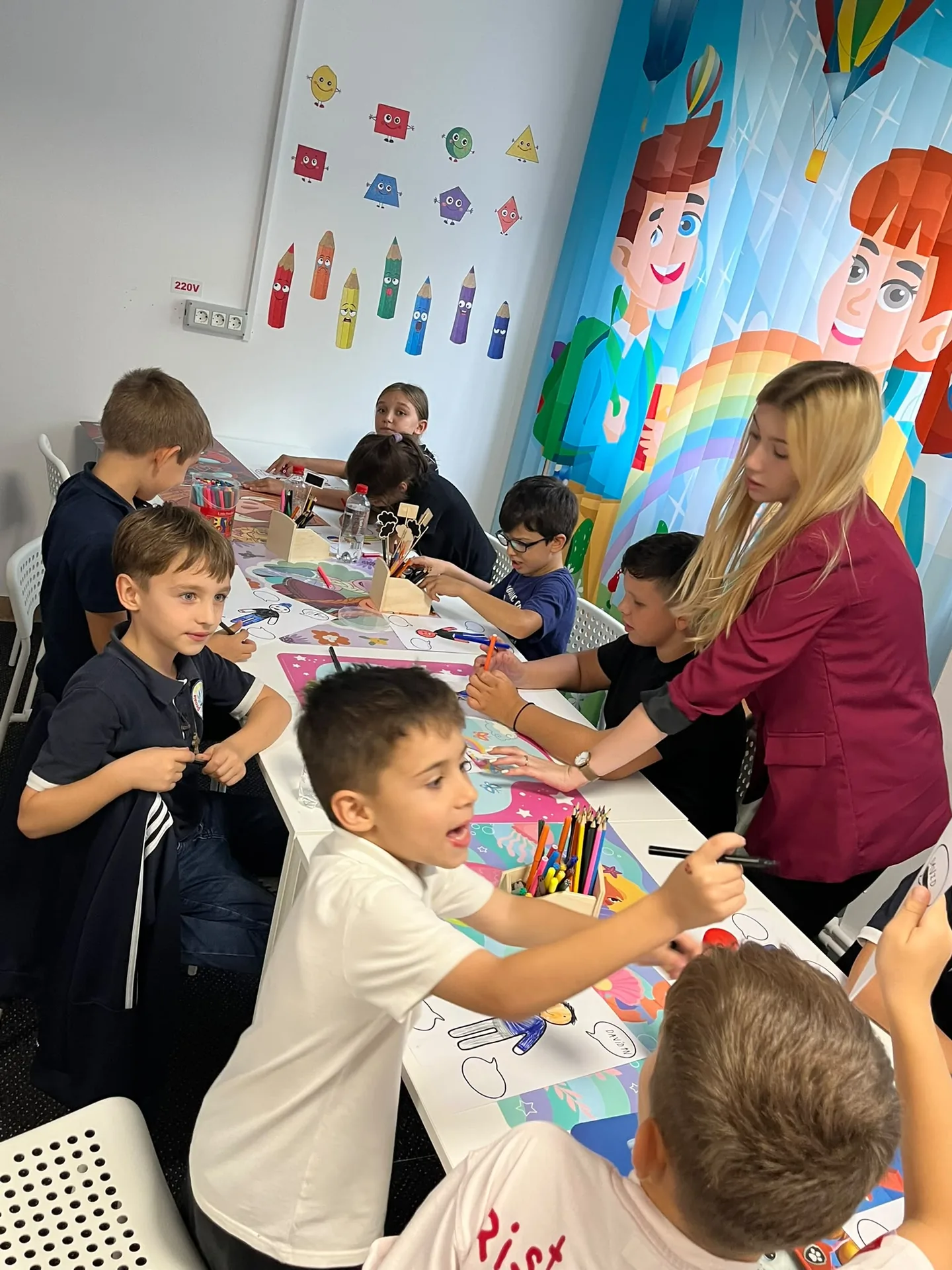 Copiii colorează la o masă cu creioane și carioci, iar o adultă stă alături de ei.