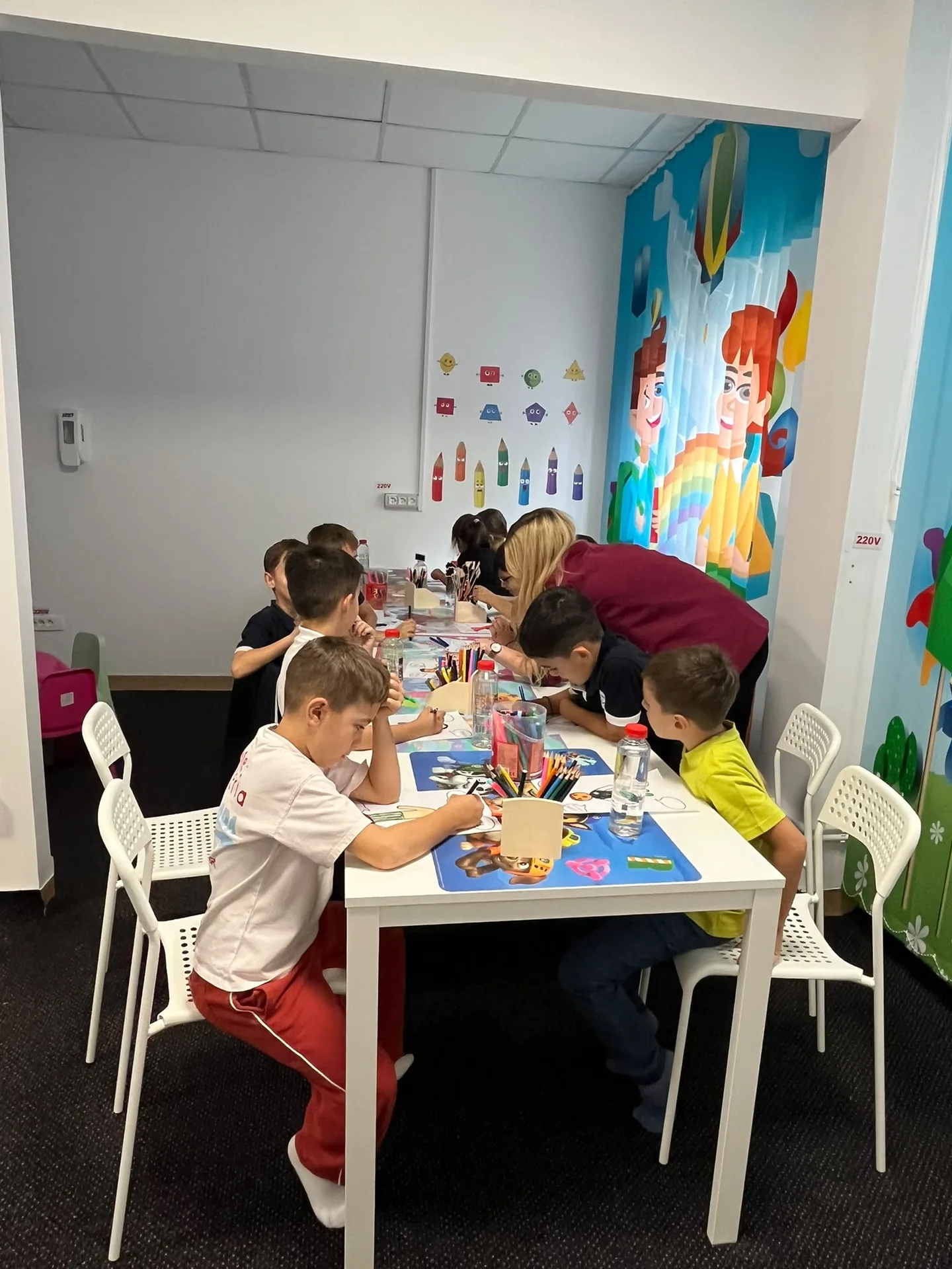 Copiii desenează la mesele albe din sala cu perdele colorate, alături de o adultă.