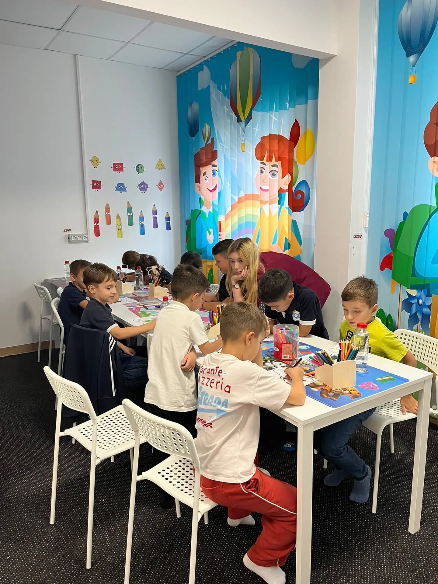 O adultă stă alături de copiii care colorează la mesele din sala de activități.