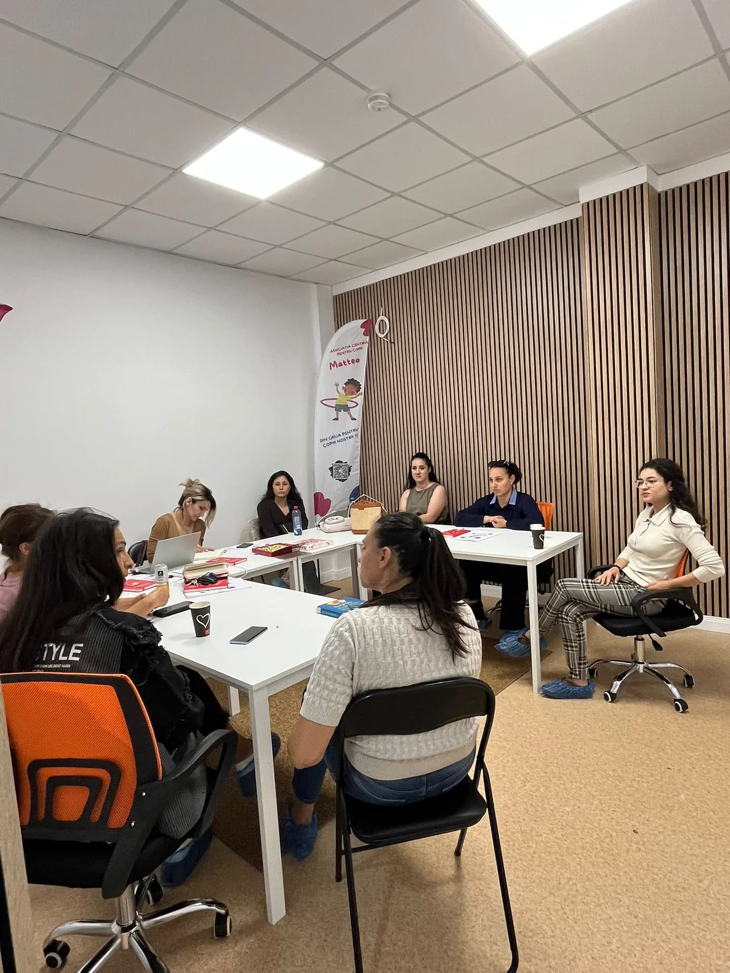 Un grup de adulți așezați în jurul meselor, într-o sală cu un perete din lamele de lemn.