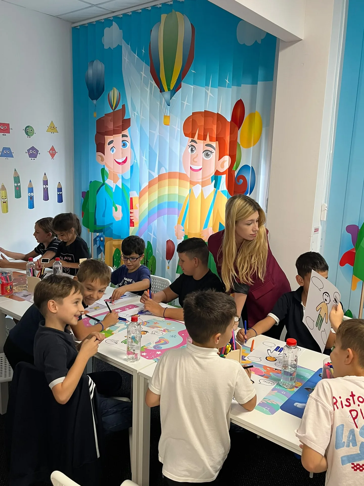 Copiii desenează împreună, iar unul dintre ei ridică o foaie colorată în dreptul feței.